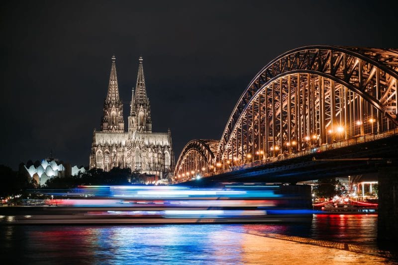 De verlichte Dom van Keulen en de Hohenzollernbrücke over de Rijn bij avond, met een passerende rondvaartboot